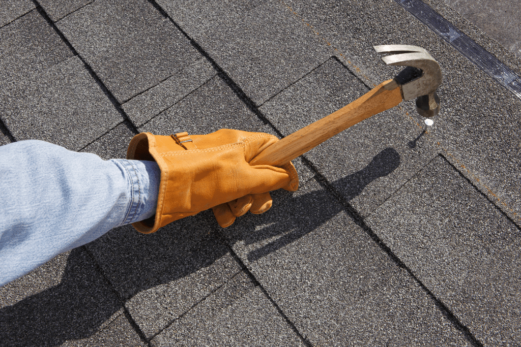 a hand with a light brown glove on holding a hammer, hammering a screw into a roof