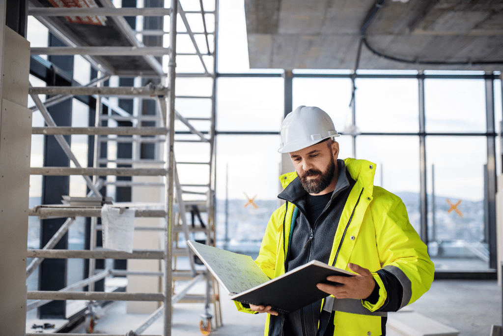 a man holding a book or folder in a building with a yellow jacket and white hard hat with scaffolding in the background